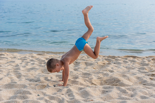 A child on the beach does sports and performs gymnastic exercises against the background of the sea.  A child does a handstand on the beach