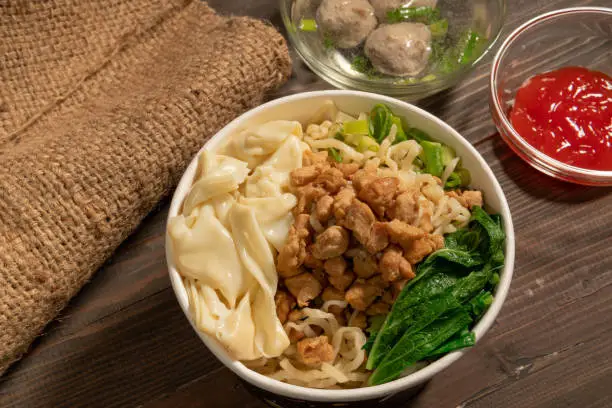 Chicken noodles in white bowl and wooden chopsticks on natural background with chicken pieces, green mustard greens, meatballs, boiled dumplings, sauce. Indonesian Chicken Noodles