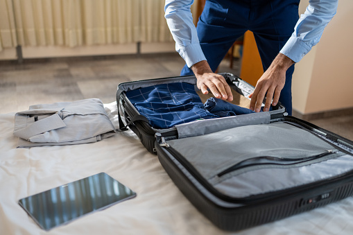 Young businessman packing suitcase, putting folded formalwear and digital tablet in baggage. Person preparing for business travel in hotel room