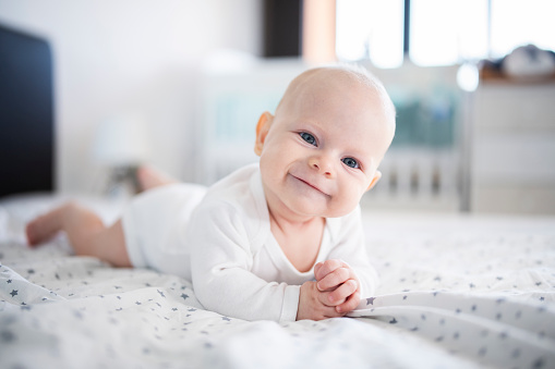 Adorable baby boy in white sunny bedroom in winter morning. Newborn child relaxing in bed. Family morning at home. Newborn kid during tummy time smiling happily at home