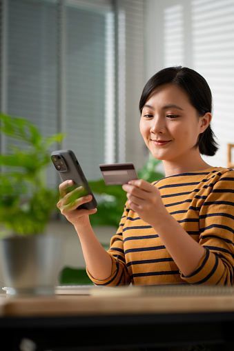 Asian woman holding credit card using smart phone for shopping online at home office, easy paying for purchase on website.