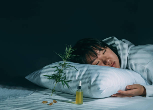 Asian girl sleeping in evening bedroom with cbd oil, capsules and a cannabis branch. Melatonin production, concept of combat sleep disorders. Dark background stock photo