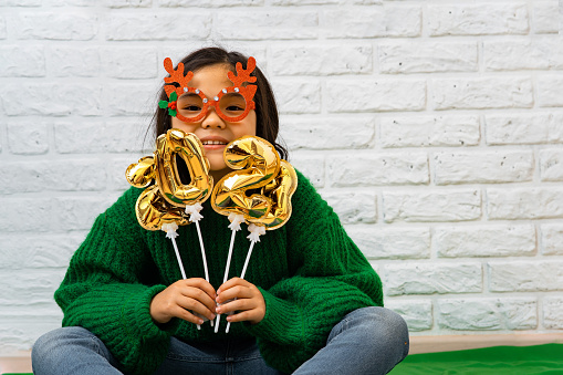 Cheerful Asian child girl in a green sweater in masquerade Christmas glasses with balls with the number 2023. Vietnamese girl in masquerade glasses with deer horns, laughs. Concept of christmas masquerade, party