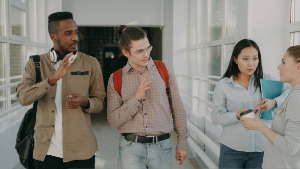 istock Two young handsome multi-ethnic friends walking down white glassy corridor in college talking passing by standing girls and waving hands to them 1434906650