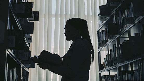 Silhouette of young student girl standing among bookshelves in big university library against window holding book turning over pages and reading