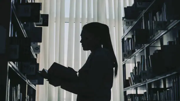 Photo of Silhouette of young student girl standing among bookshelves in big library against window holding book turning over pages and reading