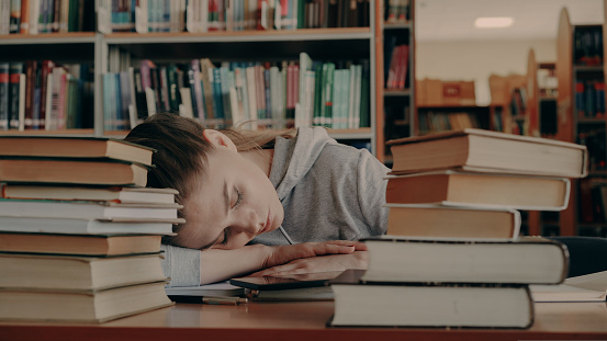 Young beautiful caucasian female student is sleeping with head on table surrounded by piles of books and papers, phone is lieing in front of her, she is in large university library