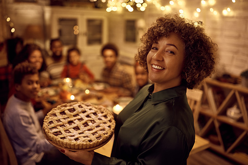 Happy mature black woman serving sweet pie to her family for Thanksgiving in dining room and looking at camera.