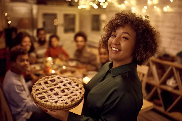 Photo of Happy mature black woman serving sweet pie to her family for Thanksgiving in dining room and looking at camera.