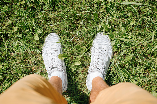 Close-up of man in shorts wearing sports stylish white summer sneakers standing on green grass lawn outdoors on sunny day, top view