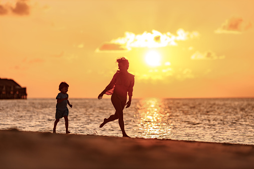 Happy mother and her small son having fun while running on the beach at sunset. Copy space.