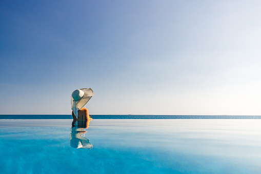 Back view of a woman with sun hat spending her summer day in infinity pool and looking at view. Copy space.
