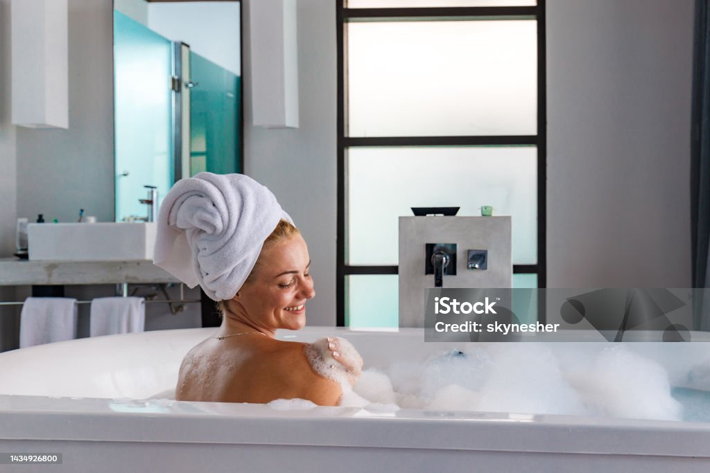 Young happy woman enjoying in bubble bath. Happy woman with hair wrapped in a towel enjoying while having a bubble bath in bathroom. Adult Stock Photo