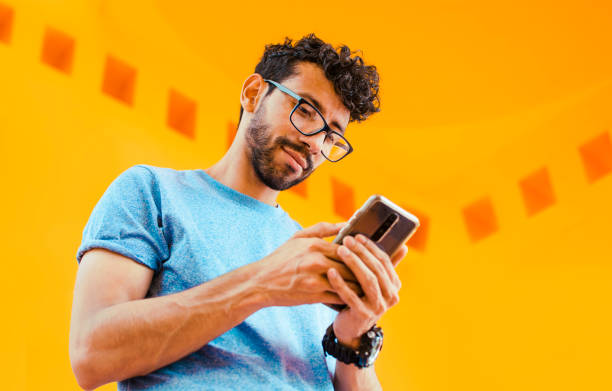 istock Low angle view of young man using cell phone outdoors. Guy sending a message with a cell phone near a yellow wall outdoors. Lifestyle of handsome bearded man using cell phone outdoors 1434926863