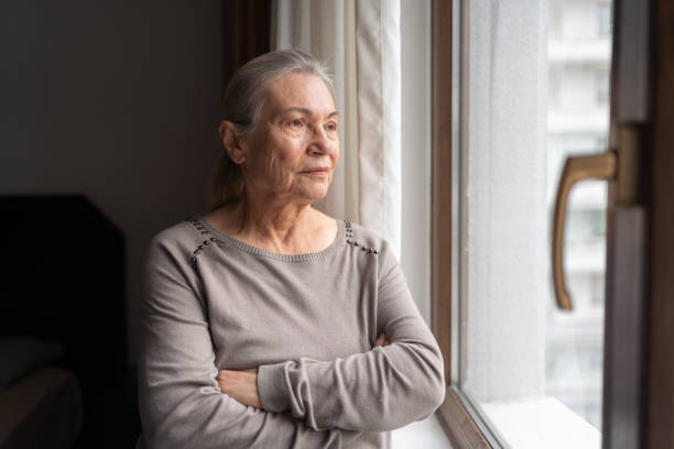 mujer mayor solitaria mirando por la ventana - pensive senior adult looking through window indoors fotografías e imágenes de stock