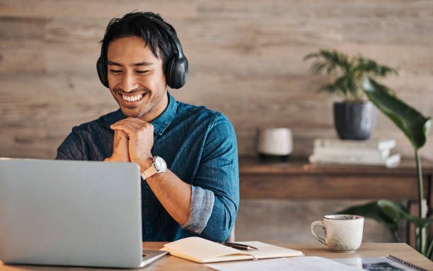 Remote worker, laptop and asian man with headphones streaming online or happy during video conference at desk with wifi. Young freelance entrepreneur working from home listening to audio music stock photo