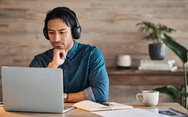 Photo of Businessman, headphones and laptop webinar in office with coffee on table, video call or watching video. Zoom call, video conference and male from Canada in online meeting with book and pen on desk.