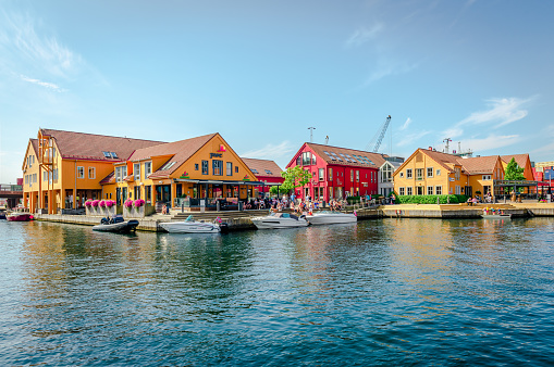 Kristiansand, Norway - August 14 2022: People enjoy a sunny day at Fiskebrygga (The Fish Wharf). Wood-fronted buildings facing the canal and boats.