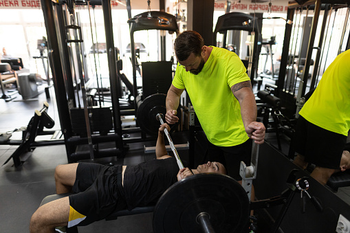 Two men, personal trainer assisting a young man in exercise on bench press in gym.