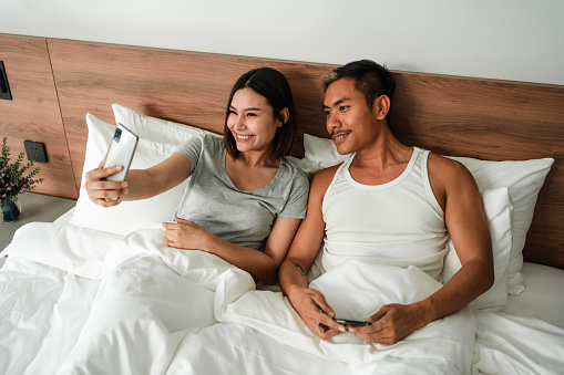 Smiling couple is taking selfies while laying in bed on their vacation in a luxury villa.