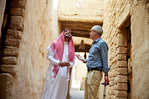 Low angle view of young man in traditional attire pausing to talk and laugh with mature Caucasian businessman as they tour al-Ula old town.