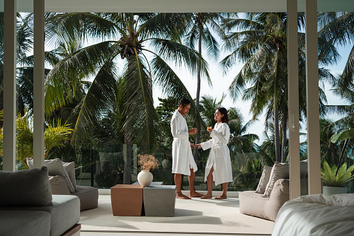 Smiling couple is standing on the balcony, holding glasses with orange juice and talking.