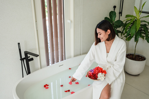 Smiling young woman is sitting on the edge of a bathtub and putting flower petals in the milk bath.