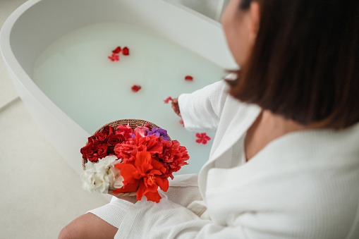 Over-the-shoulder view of a woman sitting on the edge of a bathtub and putting flower petals in the milk bath.