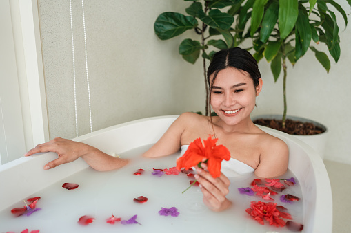 Smiling young woman is relaxing in a milk bath with flower petals.