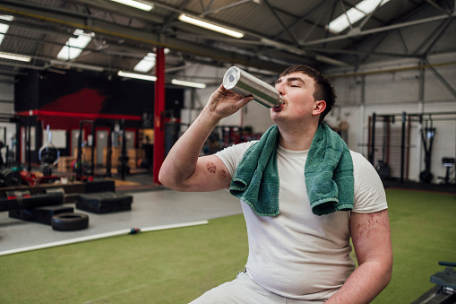 A strongman with a towel around his neck, sitting down in a gym in Newcastle upon Tyne, England surrounded by exercise equipment. He is taking a drink from a water bottle while he has a rest.