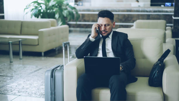 istock Tilt up of confident attractive businessman talking mobile phone and using laptop computer while sits on armchair in luxury hotel with luggage suitcase near him. Travel, business and people concept 1434955907