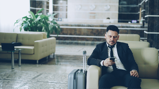 Pan shot of concentrated businessman using smartphone sitting on armchair luxury hotel with luggage near him