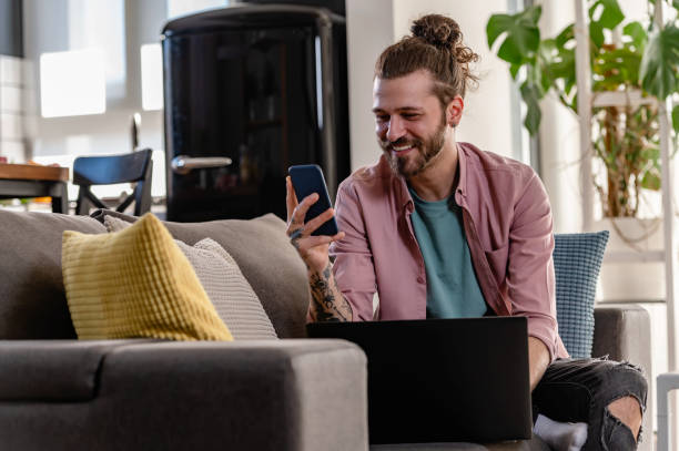 istock Young cheerful man with a sleeve tattoo using laptop computer and smartphone. Freelance entrepreneur working from home using banking apps. 1434958739