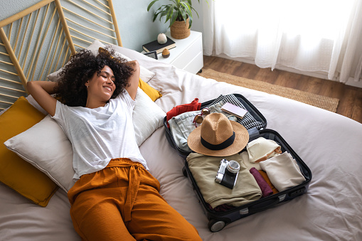 Happy multiracial woman lying down on bed next to open suitcase full of clothes, camera, passport and hat, ready to go on summer vacation trip. Holiday and vacations concept.