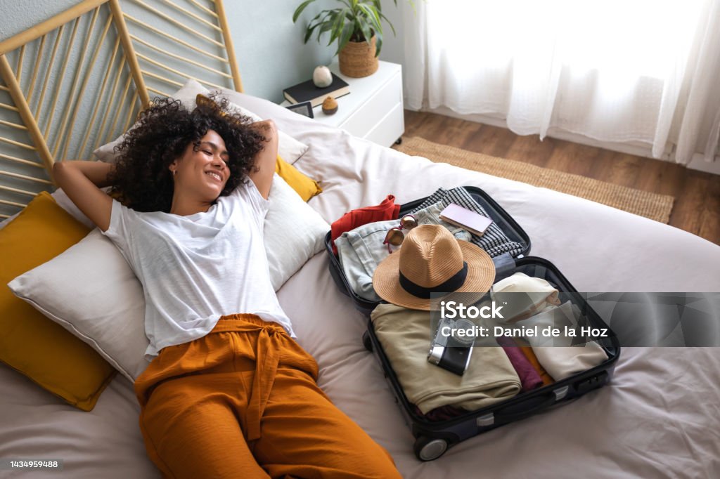 Happy woman lying down on bed next to open suitcase full of clothes, passport and hat, ready to go on vacation trip. Happy multiracial woman lying down on bed next to open suitcase full of clothes, camera, passport and hat, ready to go on summer vacation trip. Holiday and vacations concept. Vacations Stock Photo