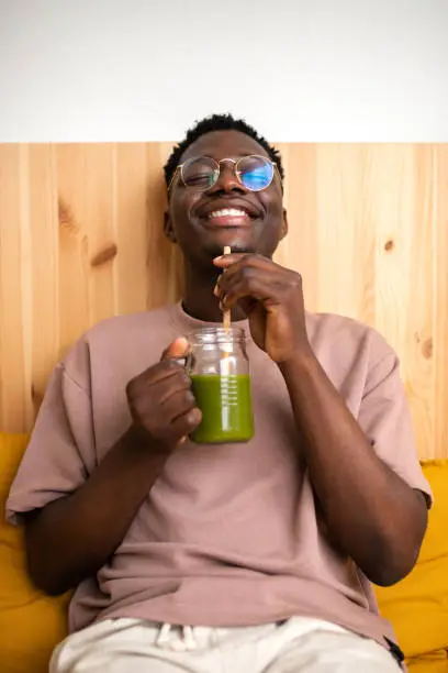 Photo of Black man drinking healthy green juice with bamboo straw at home. Vertical image.