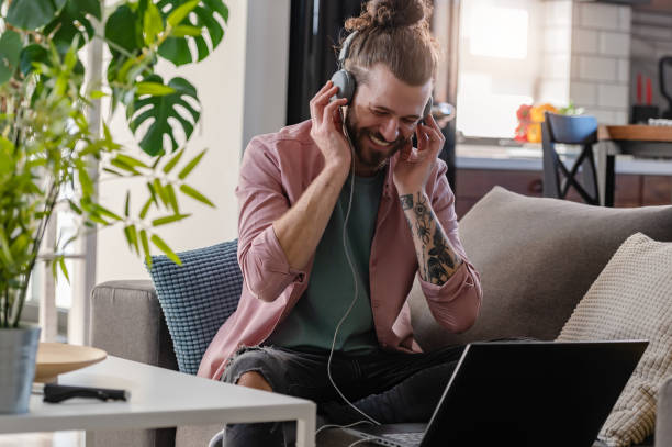 istock Young cheerful man with a sleeve tattoo using a laptop computer and smartphone listening to music online over headphones 1434959616