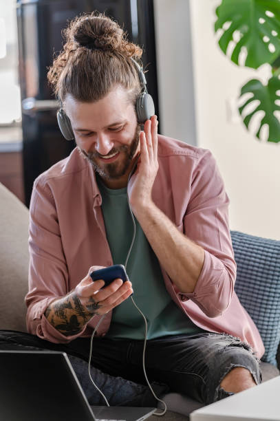istock Young cheerful man with a sleeve tattoo using a laptop computer and smartphone listening to music online over headphones 1434959967