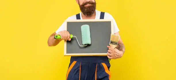 cropped man housepainter hold paint roller brush and blackboard with copy space on yellow background.