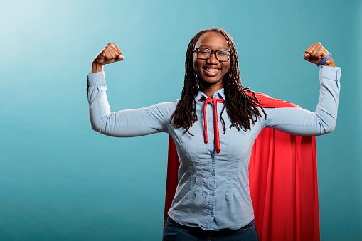 Proud and tough looking young adult superhero woman showing empowerement and braveness. Happy positive justice defender flexing arms as a sign of strength while standing on blue background.