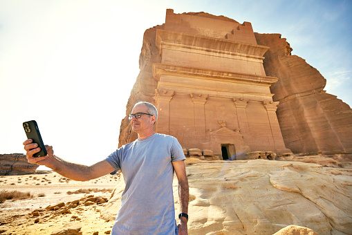 Waist-up view of mature Caucasian man holding smart phone and capturing memory with 1st-century CE sandstone architecture in background.