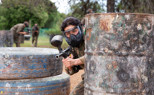 Asian female paintball player in protective mask and with gun on paintball field