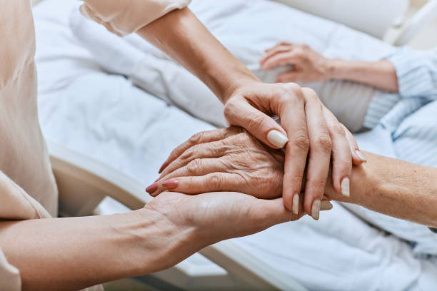 Relative holding trembling hand of senior woman with Parkinson's disease lying in hospital bed at medical ward. Diagnosis and treatment of Parkinson's disease and dementia Relative holding trembling hand of senior woman with Parkinson's disease lying in hospital bed at medical ward. Diagnosis and treatment of Parkinson's disease and dementia body care stock pictures, royalty-free photos & images