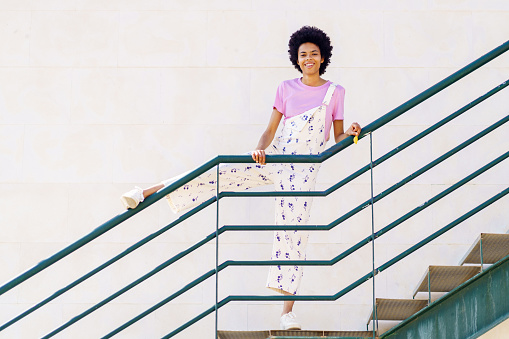 Full body of smiling African American female in casual clothes looking at camera while standing on staircase with leg on railing against white background