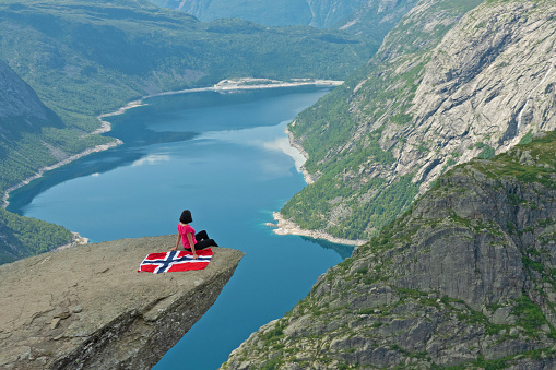 Norway - August 29, 2022: Girl sitting on Trolltunga rock with Norwegian flag. Mountain lake Ringedalsvatnet landscape.