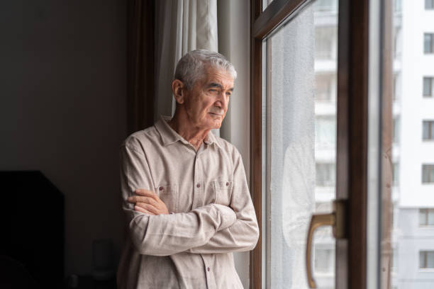 anciano solitario mirando por la ventana - pensive senior adult looking through window indoors fotografías e imágenes de stock