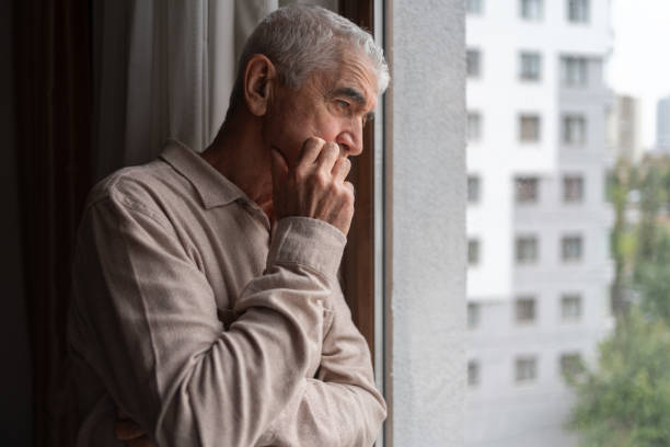 lonely senior man looking through the window - portrait men senior adult depression imagens e fotografias de stock