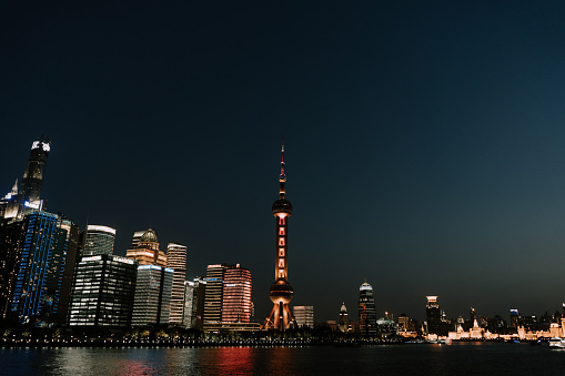 A wide shot of the city side view of Shanghai city in China at night or evening, the Shanghai World Financial Center and the Oriental Pearl TV Tower, an urban skyline including many skyscrapers or tall commercial or financial buildings, tower, shopping malls and other architectures lit up with city lights, next to the sea with boats and ships sailing on the sea, showing the lifestyle, nightlife and culture of a modern developed China city with its illuminated view.