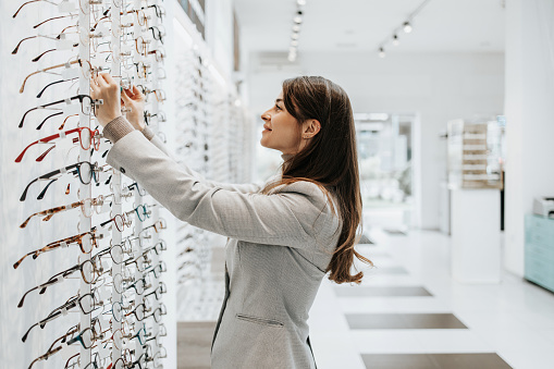 Beautiful and fashionable woman choosing eyeglasses frame in modern optical store.