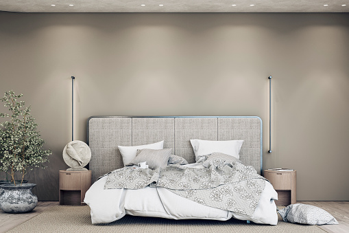 Luxury bedroom with a gray textile headboard bed, white and gray sheets - bedding,  night tables, a sculpture, pillows, a beige carpet, and vertical wall lights on the hardwood floor. A large eucalyptus plant in a pot in front of a rounded taupe (dark gray brown) empty plaster wall with copy space and built-in reflector lights on the ceiling. 3D rendered image.